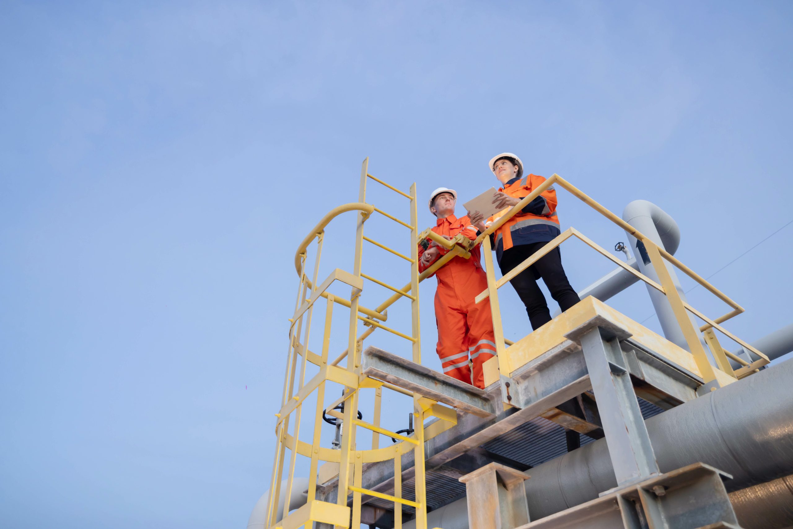 Two workers in safety gear standing on an industrial platform with large grey pipes in the background.
