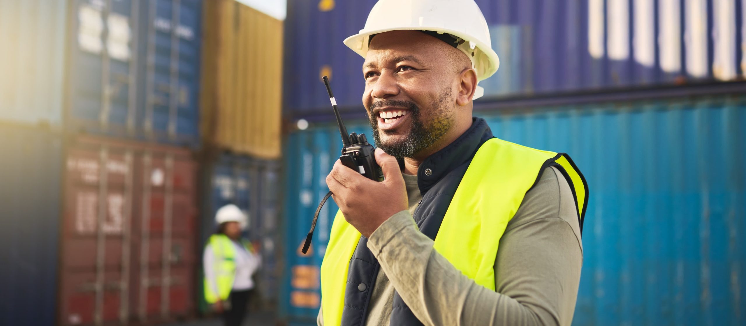 Person in a hard hat and high-visibility vest smiling with a walkie-talkie in front of shipping containers.