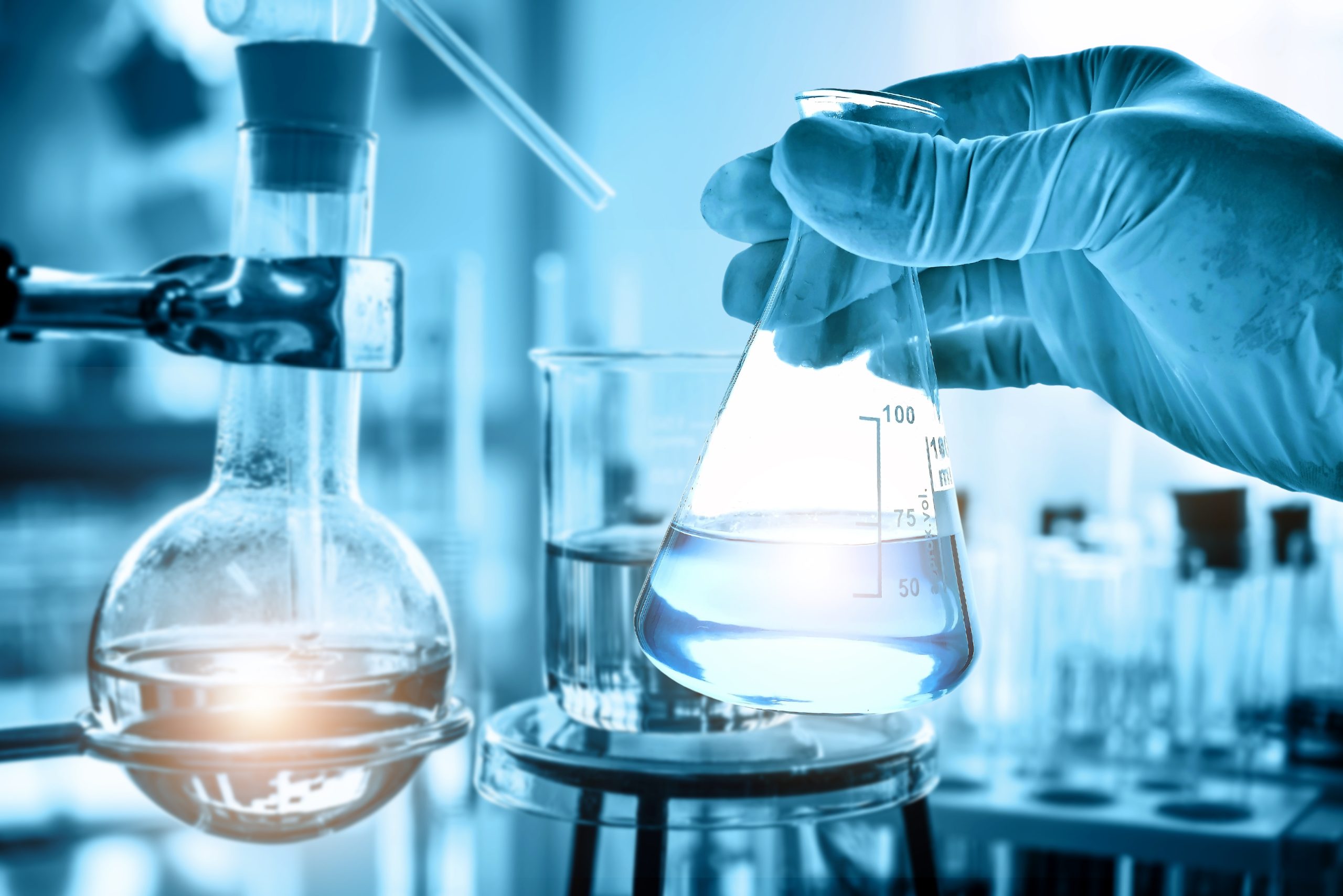 A gloved hand holding an Erlenmeyer flask with blue liquid in a laboratory.