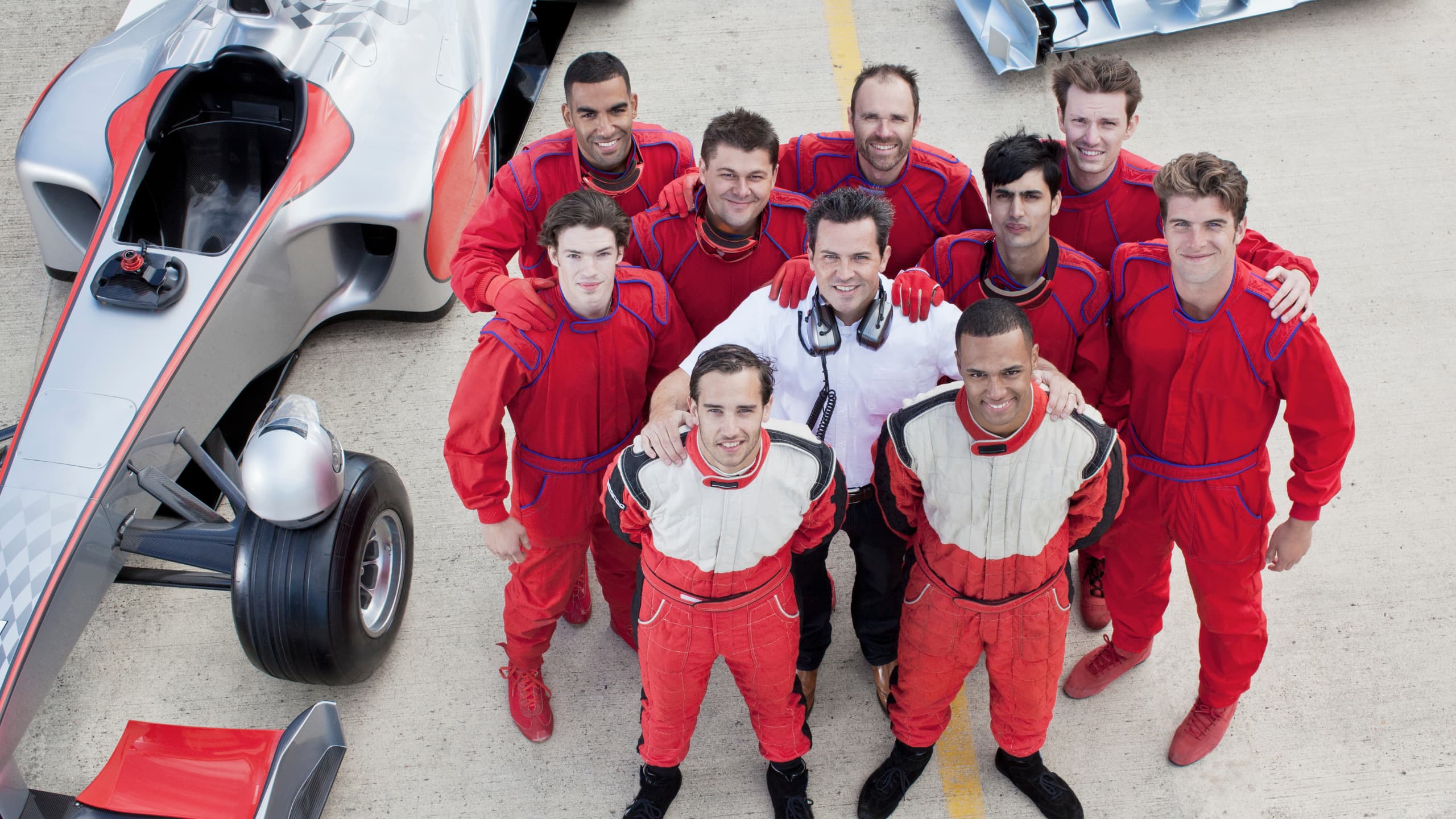 Group of men in racing suits standing beside a silver and red race car on a concrete surface.