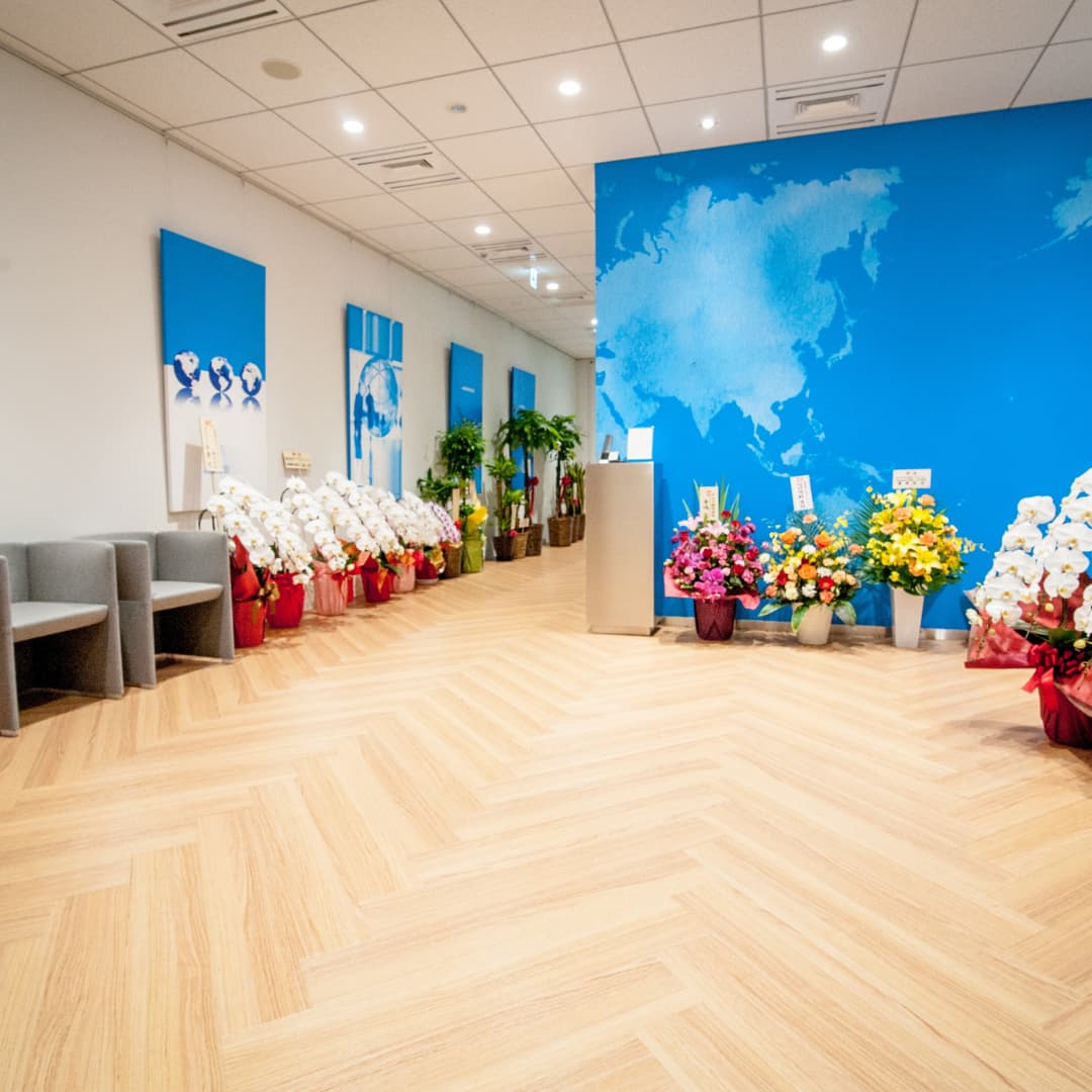 A modern lobby with light wood-patterned flooring, decorated with potted flowers and plants, blue wall panels with graphics, and a world map design.