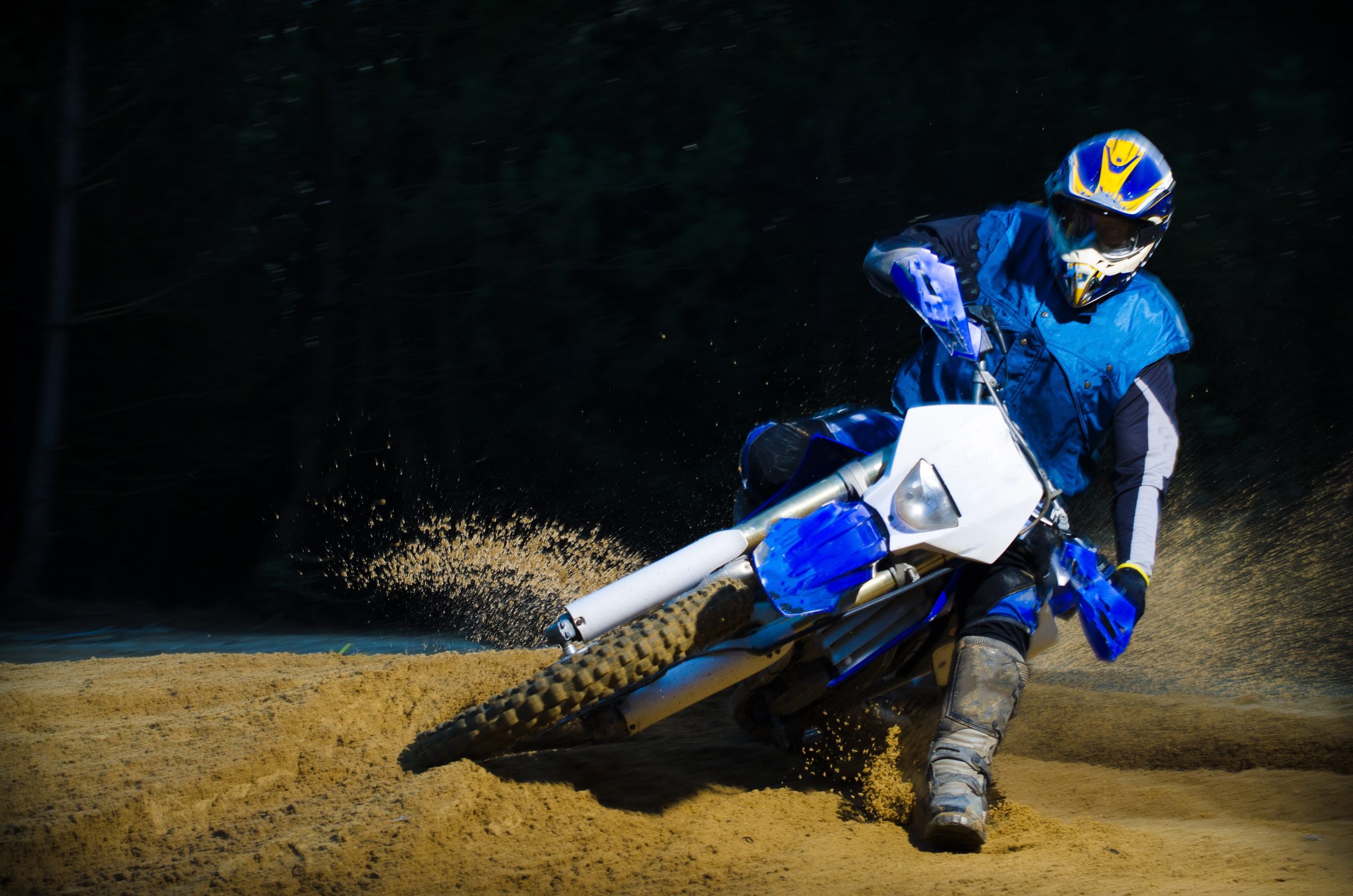 A person on a dirt bike leans into a turn on a sandy track, causing sand to spray up behind.