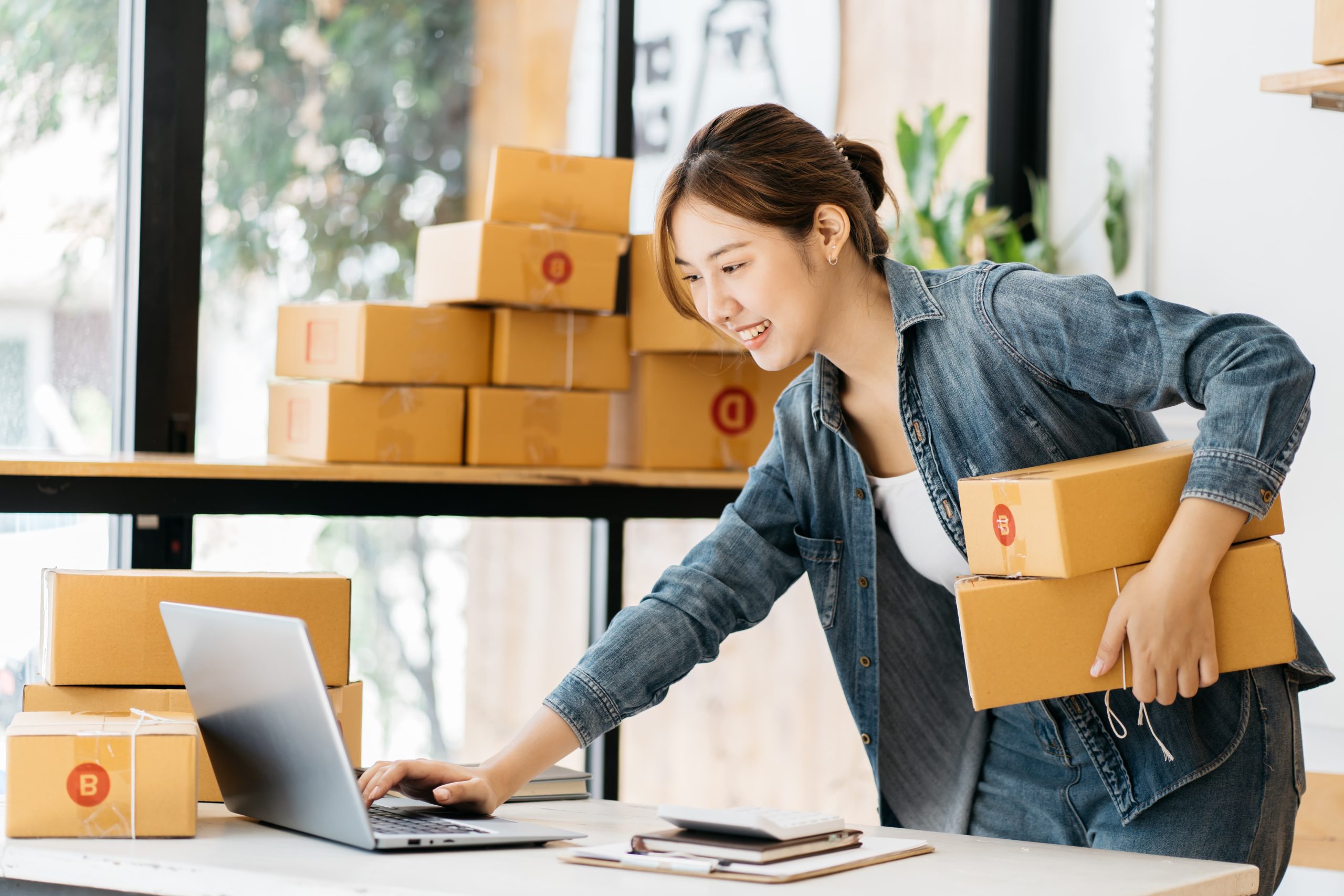 A woman in denim holds packages while using a laptop in a bright room.
