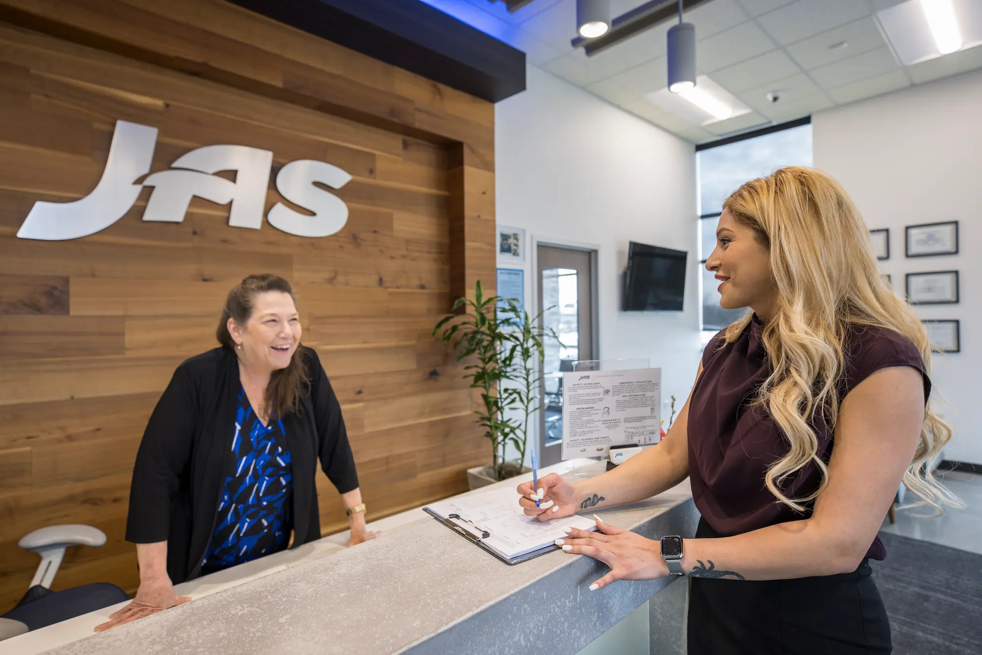 Two women engaging in a friendly conversation at a reception desk inside a JAS Worldwide office. One woman, wearing a black jacket and blue patterned blouse, leans on the desk, smiling. The other, with long blonde hair and a maroon blouse, holds a clipboard and pen while smiling back. The background features a wooden wall with the JAS logo, indoor plants, and a modern office design.