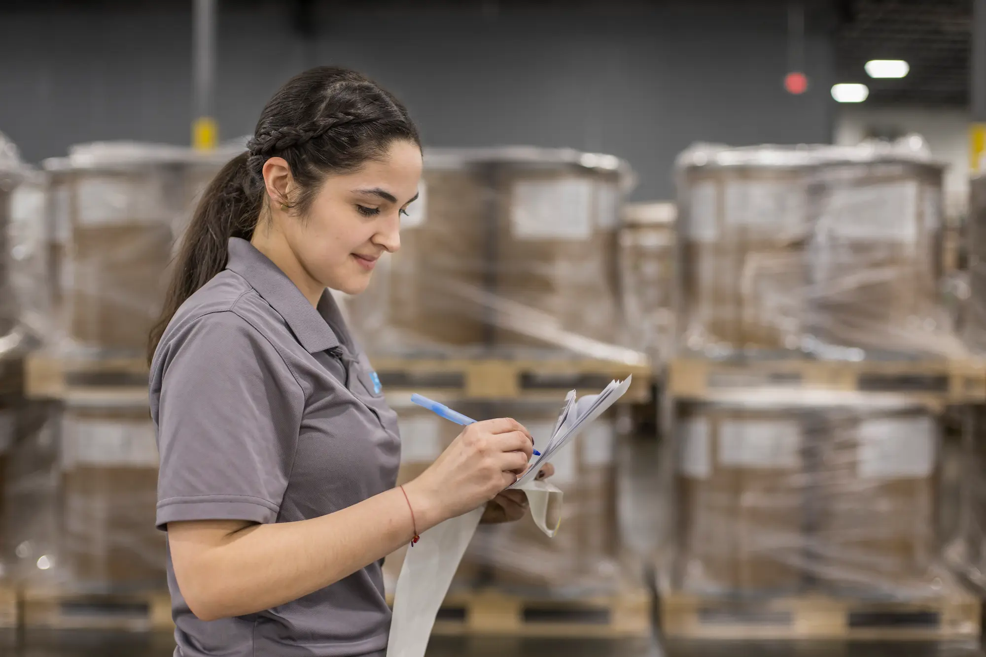 A woman in a gray polo shirt standing in a warehouse, writing on a clipboard with a pen. She is focused on her task, with rows of wrapped pallets stacked in the background, creating an organized logistics setting.