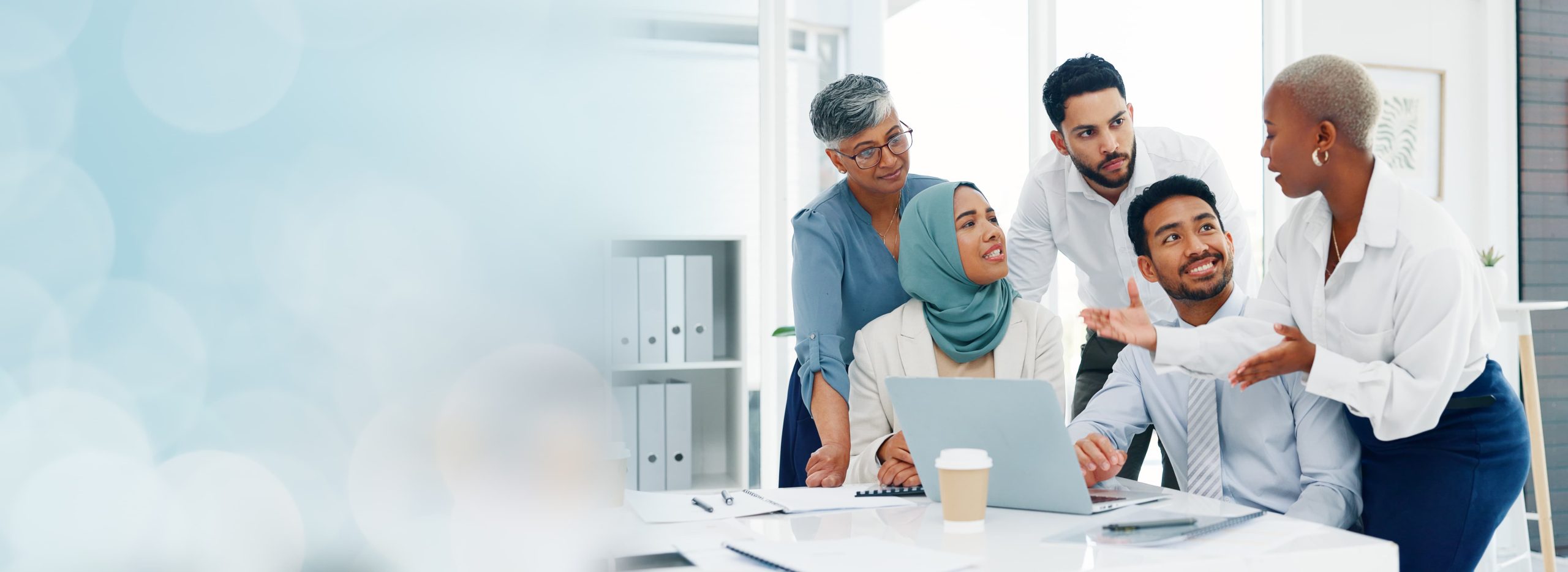 A group of five colleagues discussing around a table in an office setting with one using a laptop.