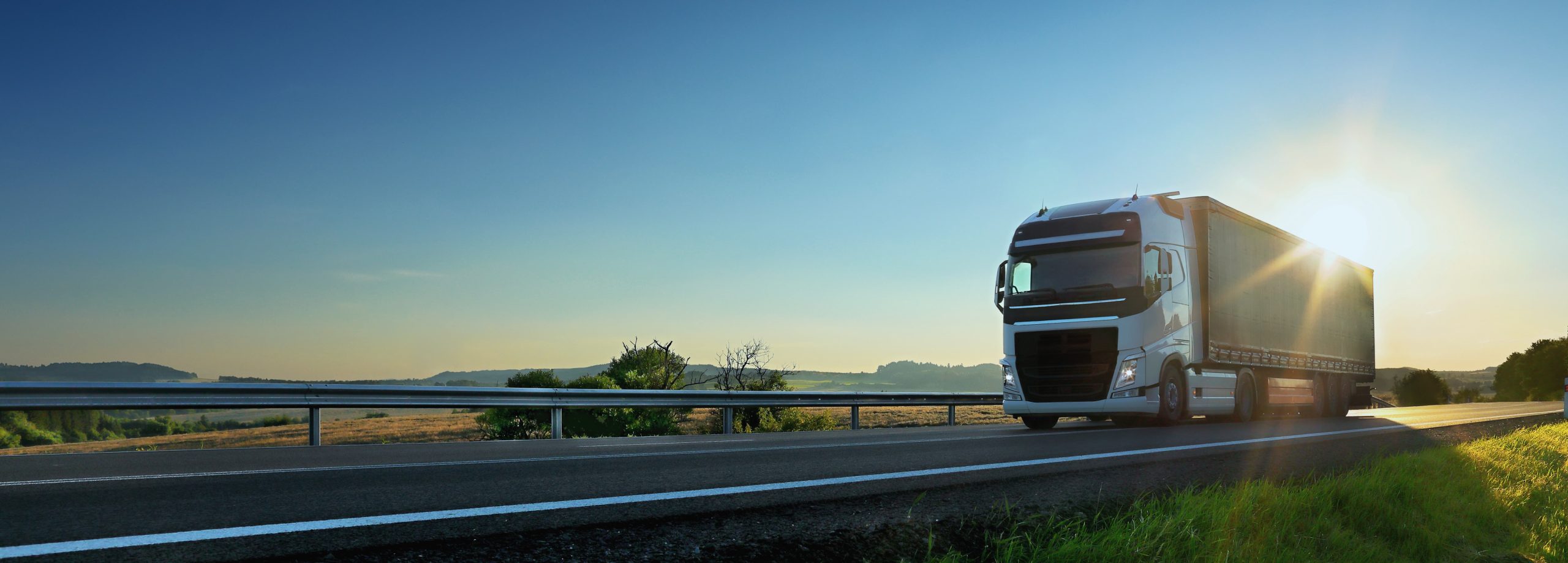 A white semi-truck drives on a highway under a clear blue sky with the sun shining brightly.