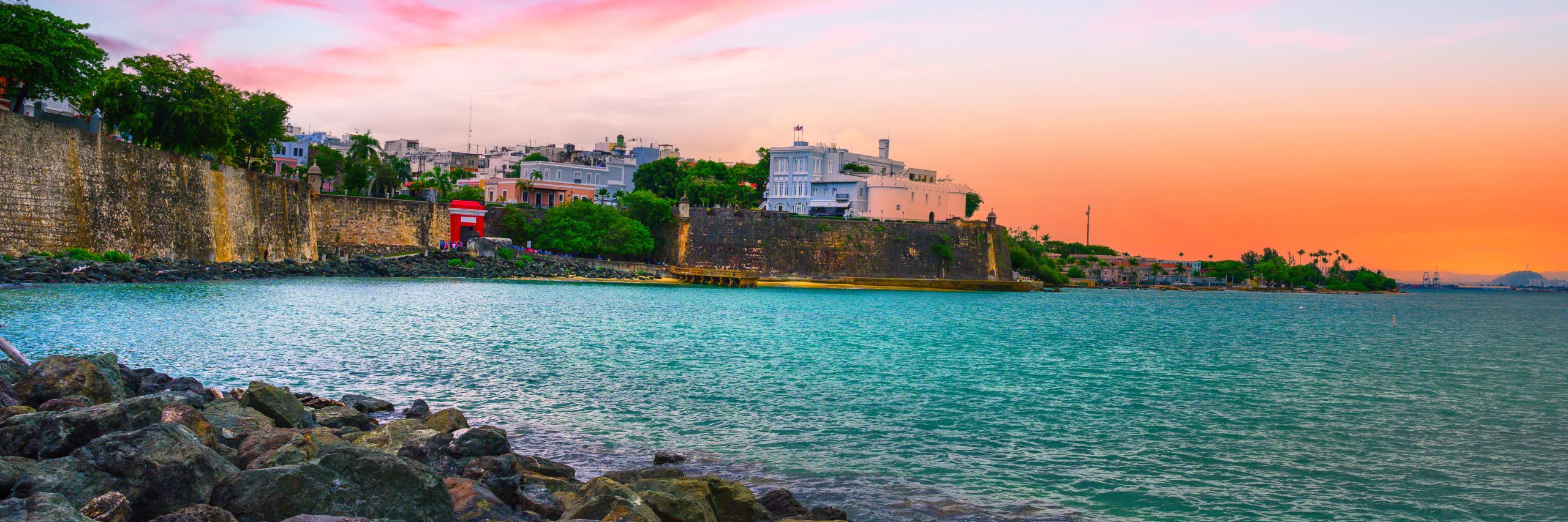 A coastal scene at sunset with a historic stone wall, red structure, city buildings, and a colorful sky.