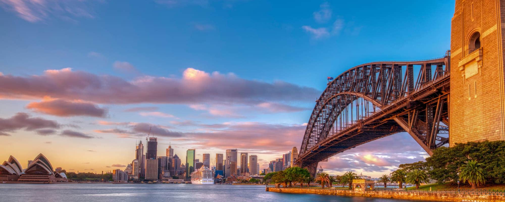 Sydney's skyline at sunset with the Opera House, Harbour Bridge, and a cruise ship in the harbor.