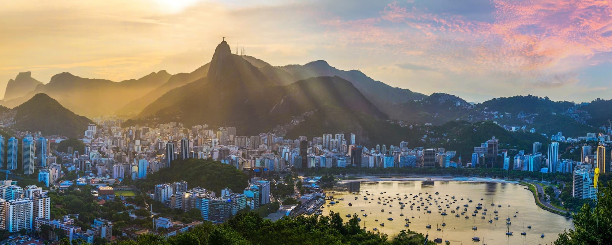 Panoramic cityscape at sunset with a mountain, statue, and a bay filled with boats.