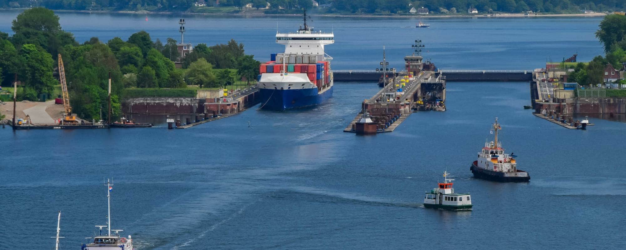 Large cargo ship in a canal with tugboats and greenery, passing through a lock system.