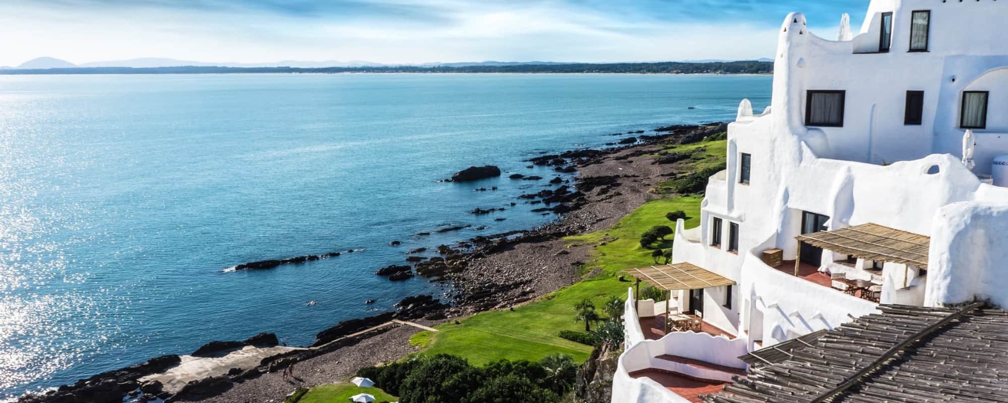 A white stucco building with curved architecture overlooking a turquoise ocean and rocky coastline.