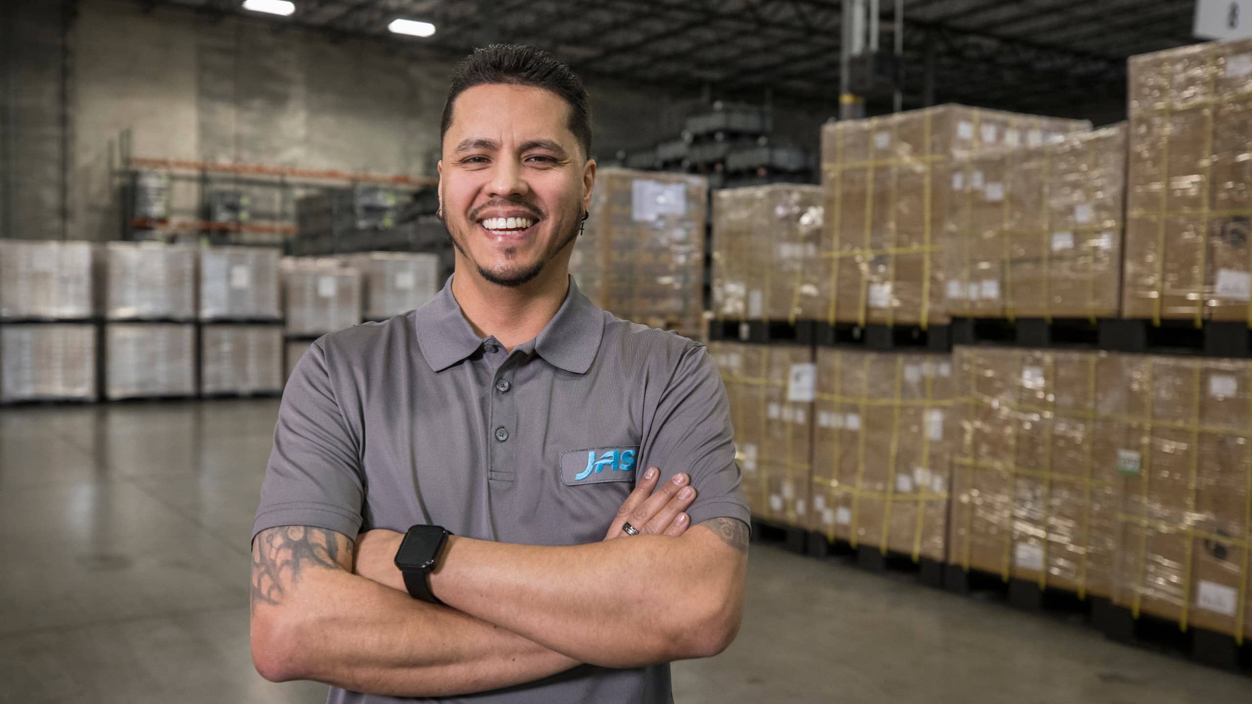Man in a gray polo shirt standing in a warehouse with stacks of cardboard boxes.