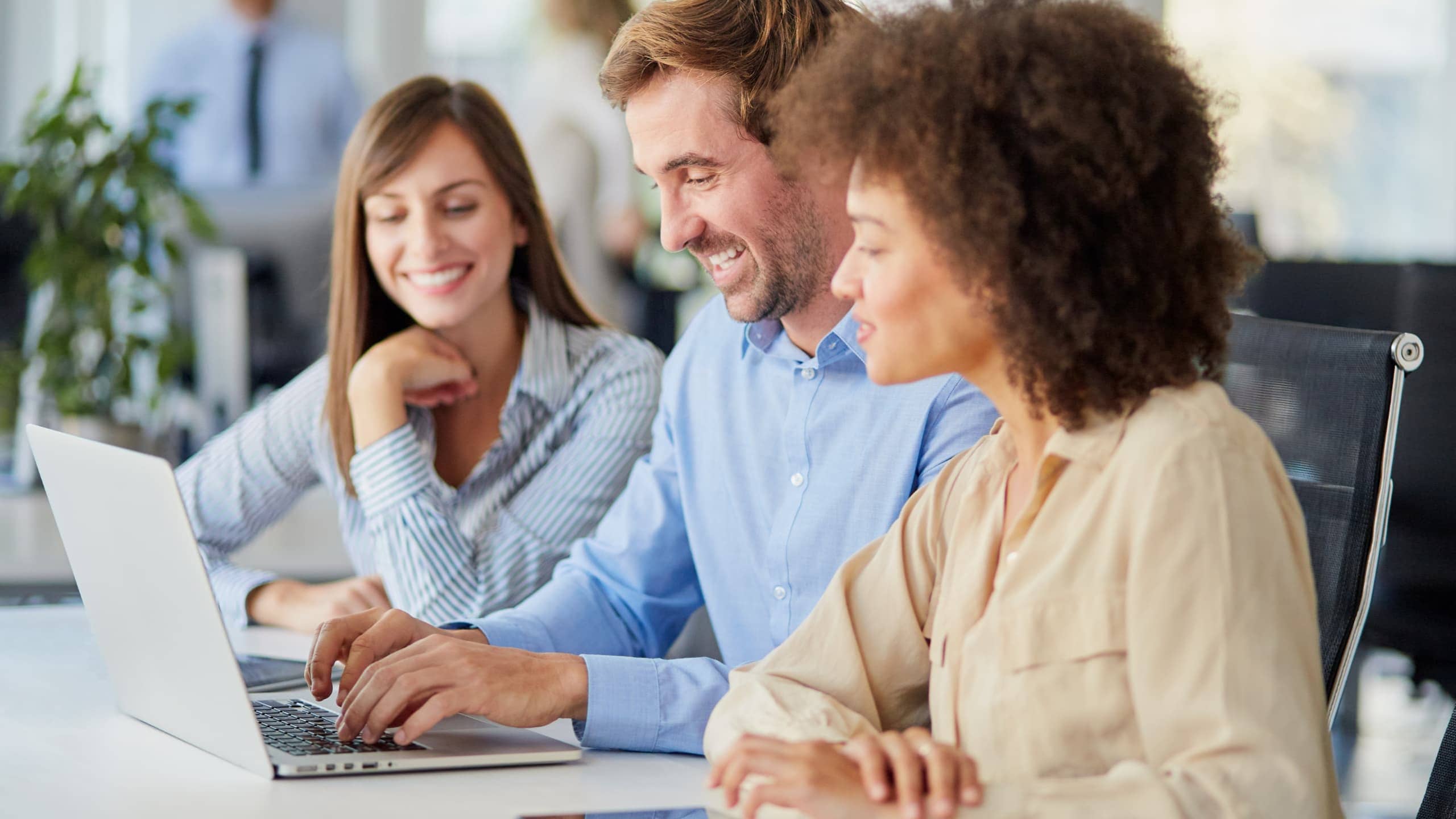 Three colleagues smiling and working together on a laptop in an office setting.