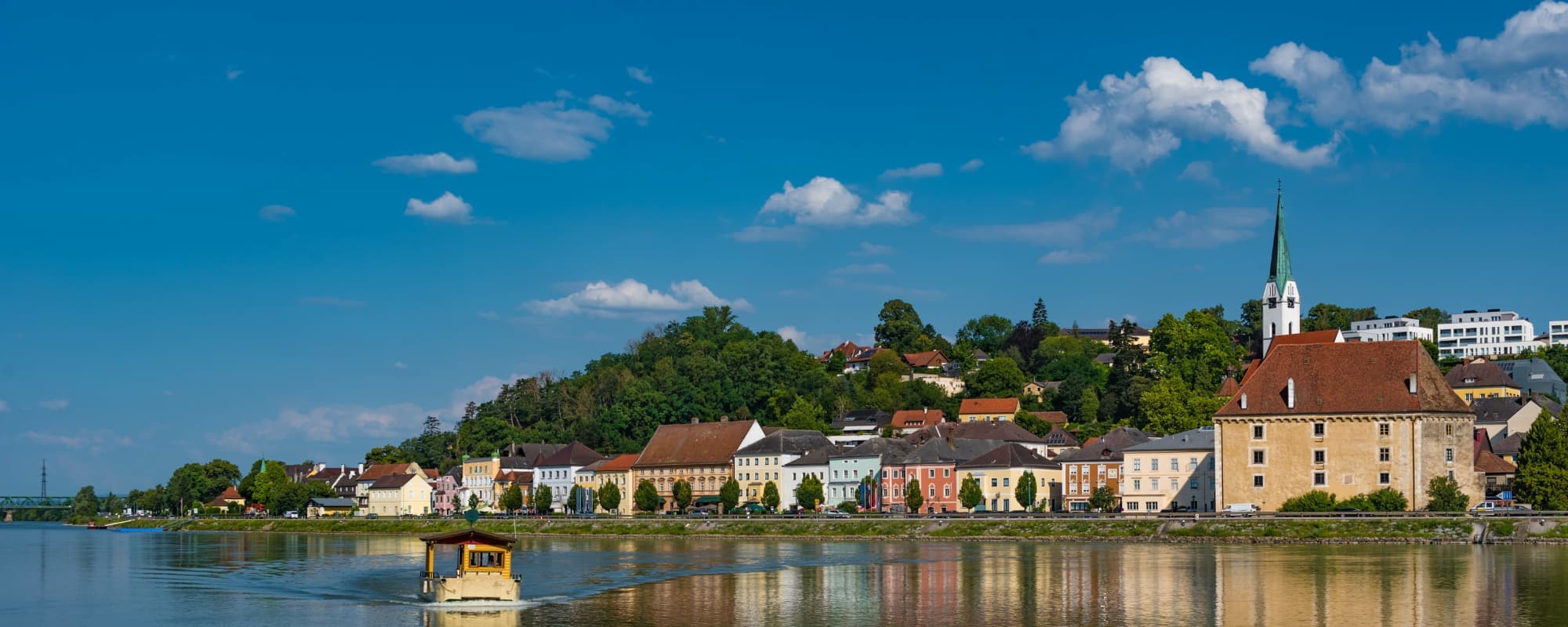Scenic view of a colorful riverside town with historic buildings, a church spire, and a green hillside under a blue sky.