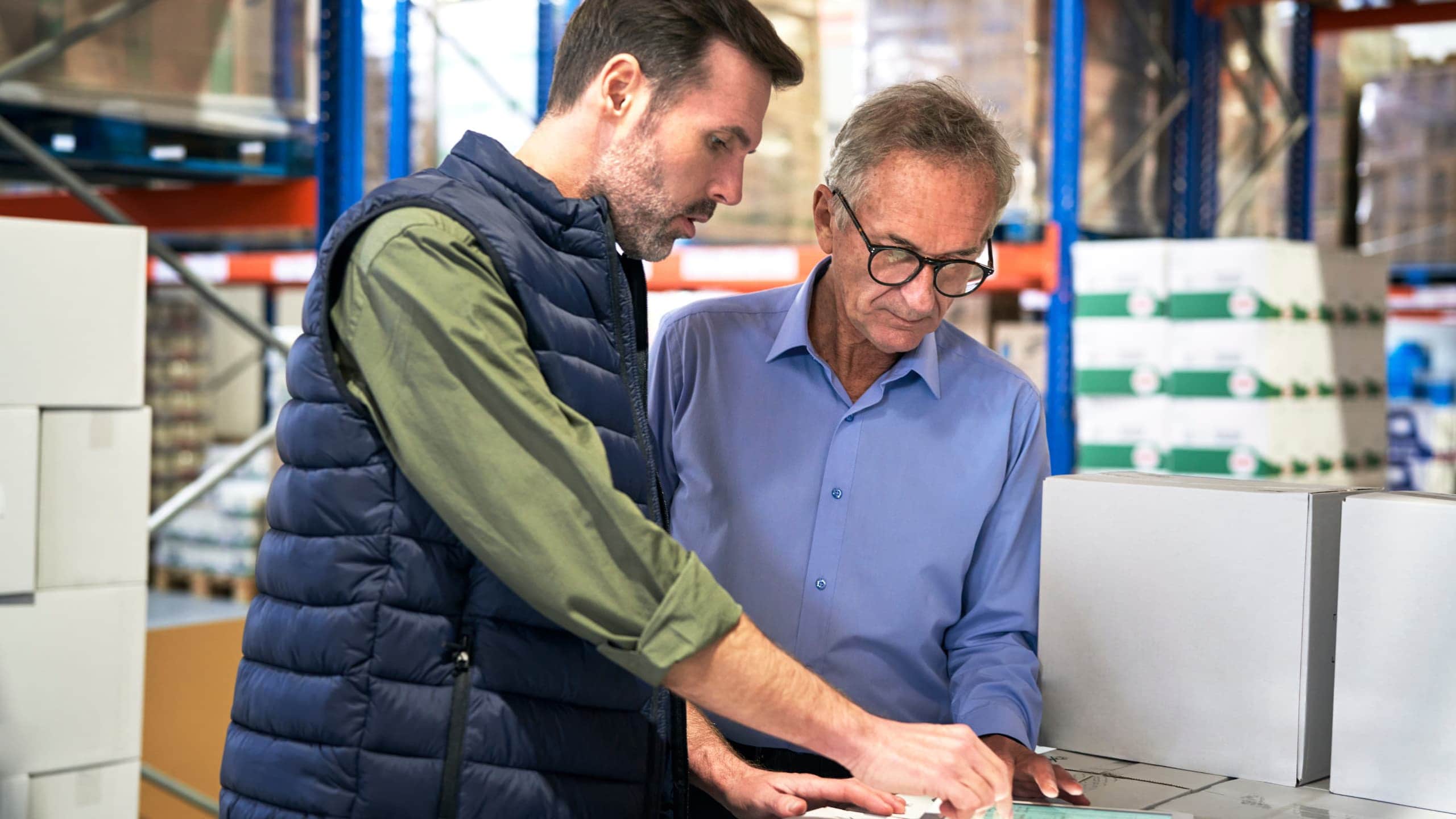 Two men discussing a document in a warehouse near stacked boxes.