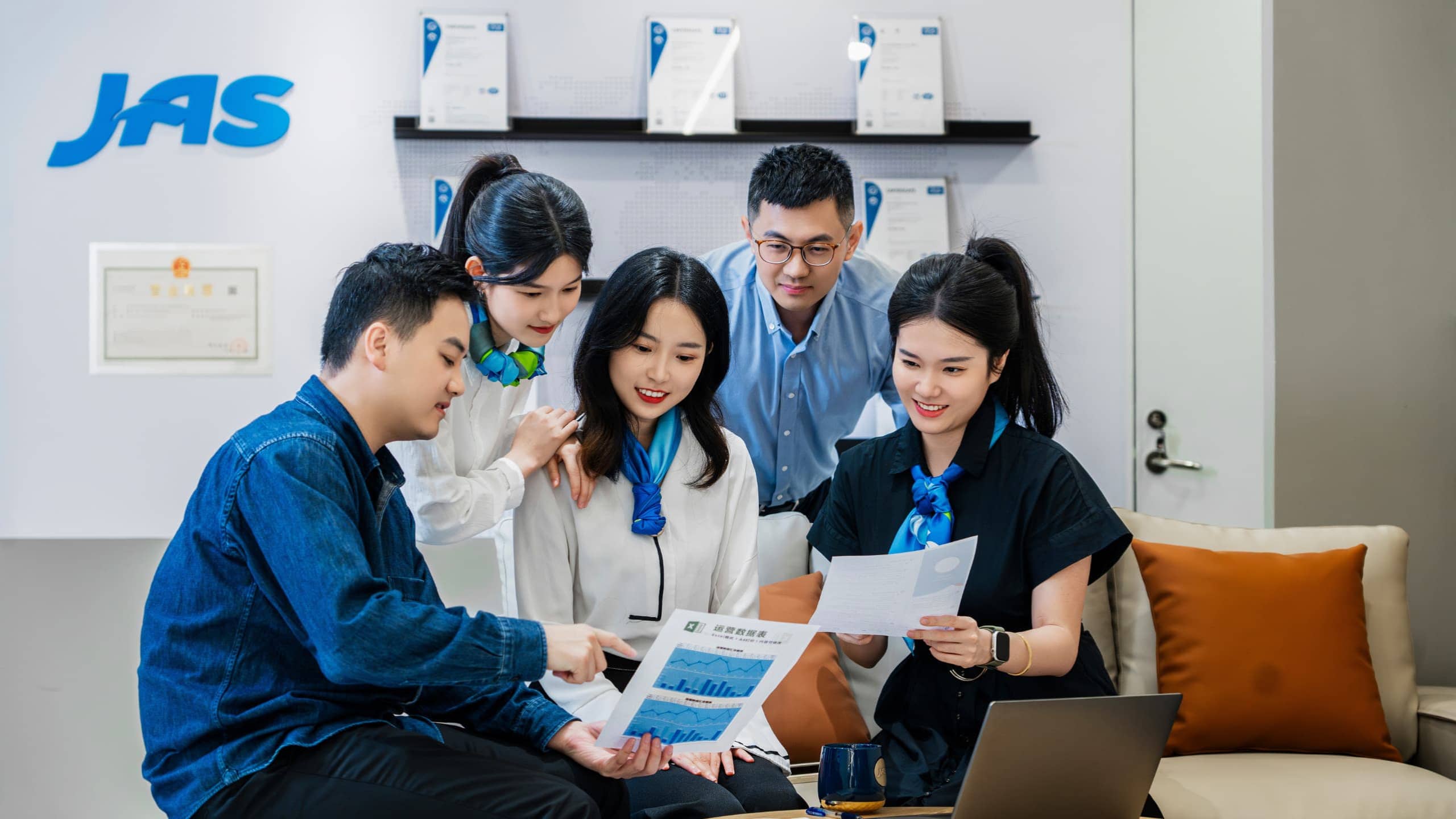A group of five people in an office discussing documents, with the JAS logo and certificates on the wall behind them.