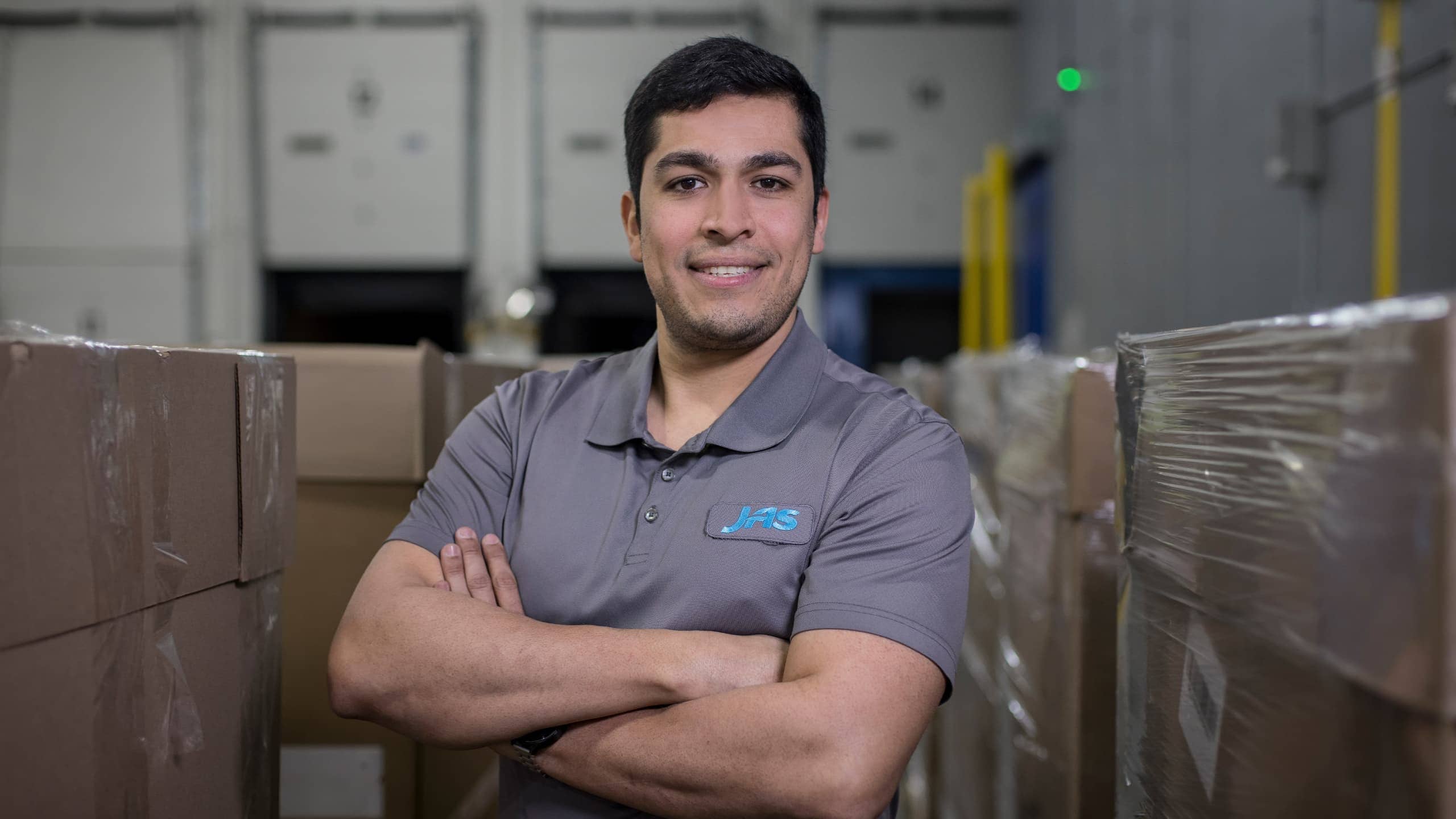 A person smiling with crossed arms in a warehouse filled with stacked cardboard boxes.