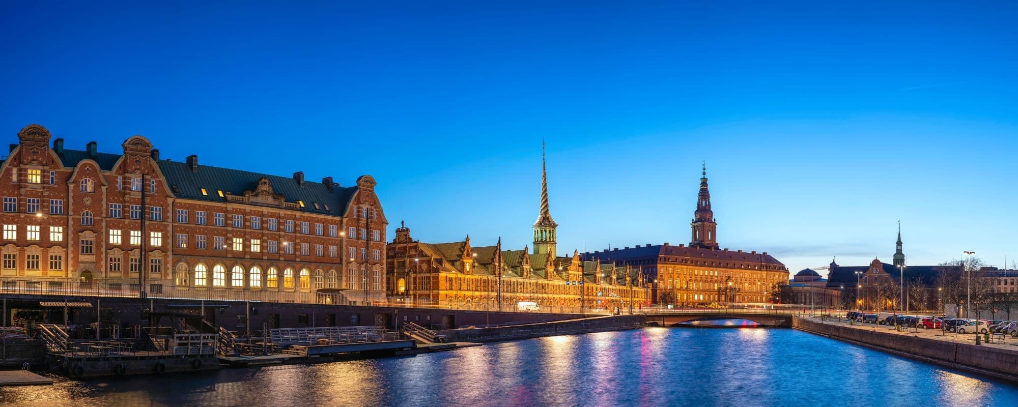 Panoramic evening view of a cityscape along a canal with historic buildings and distinctive spires.