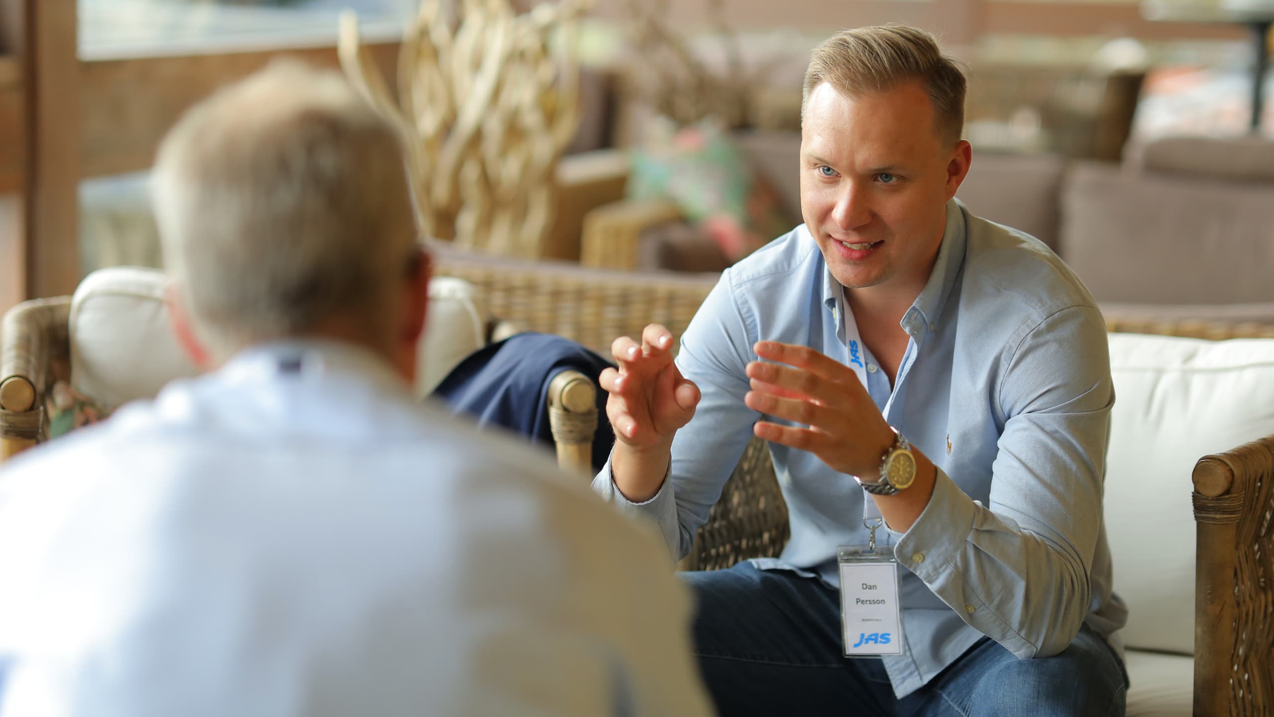 Two people seated in conversation, focusing on a man in a blue shirt gesturing.