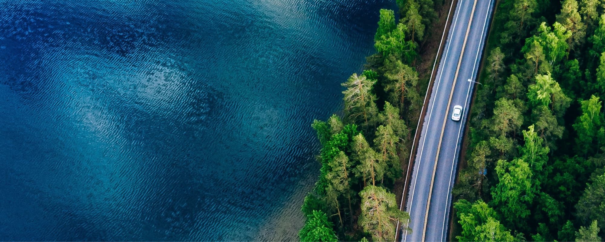 Aerial view of a road with a white car beside a blue lake and surrounded by dense green forest.