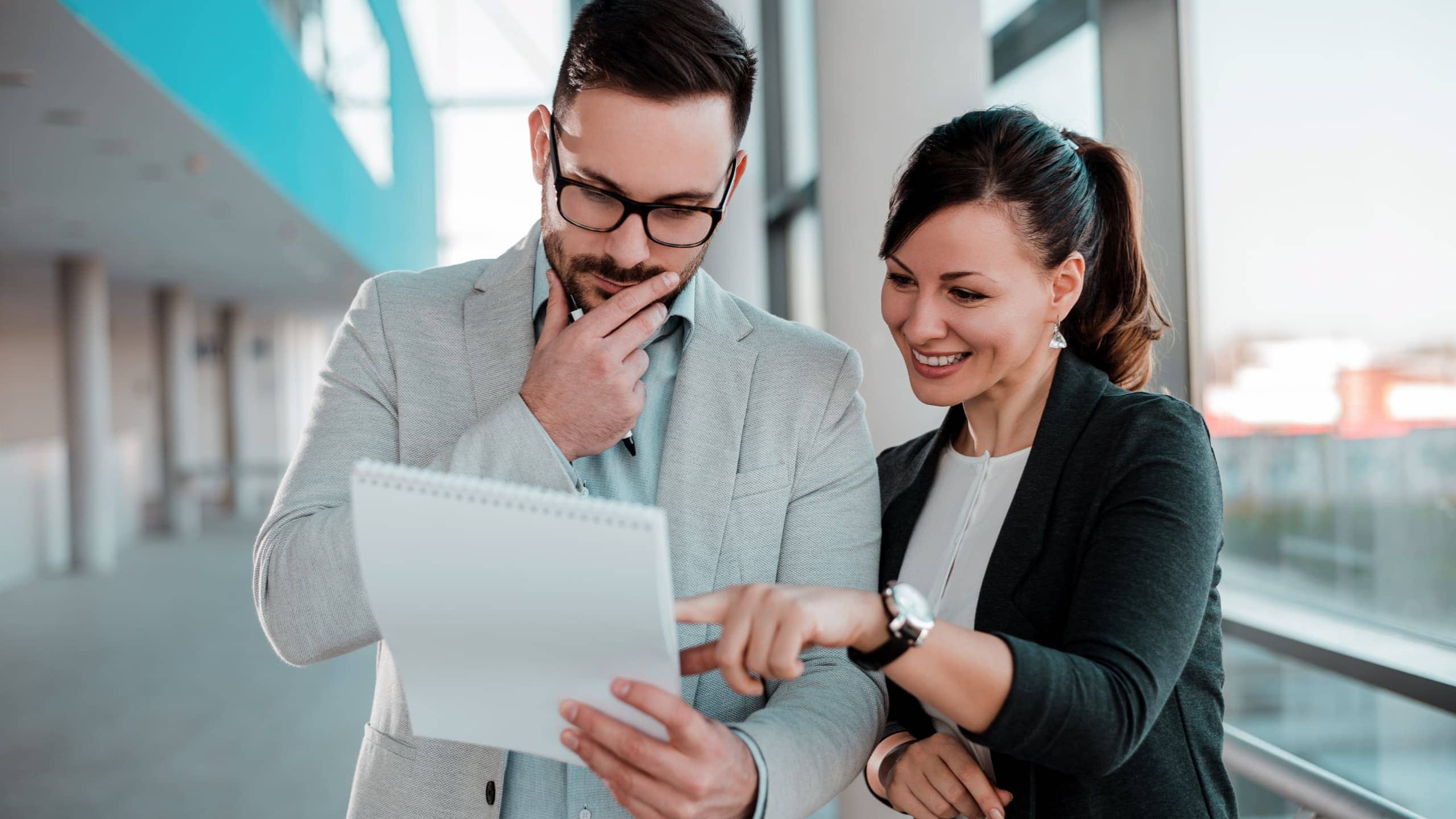 Two people in business attire examine a notepad in a bright hallway.