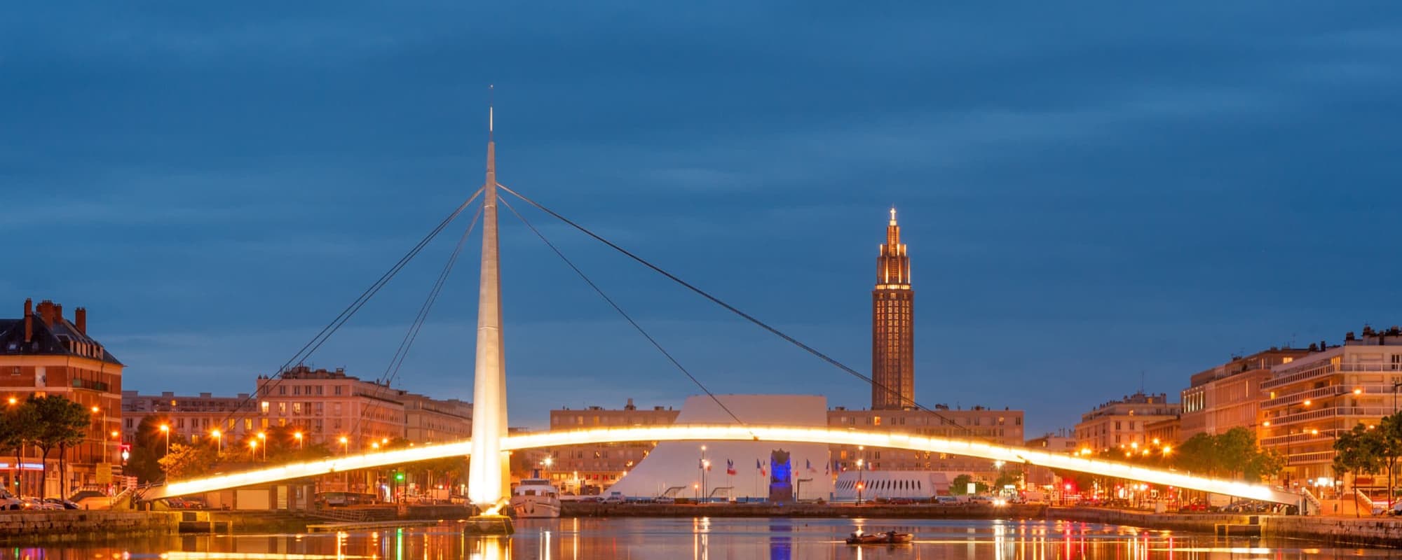 Modern illuminated suspension bridge over water at twilight with a tall building in the background.