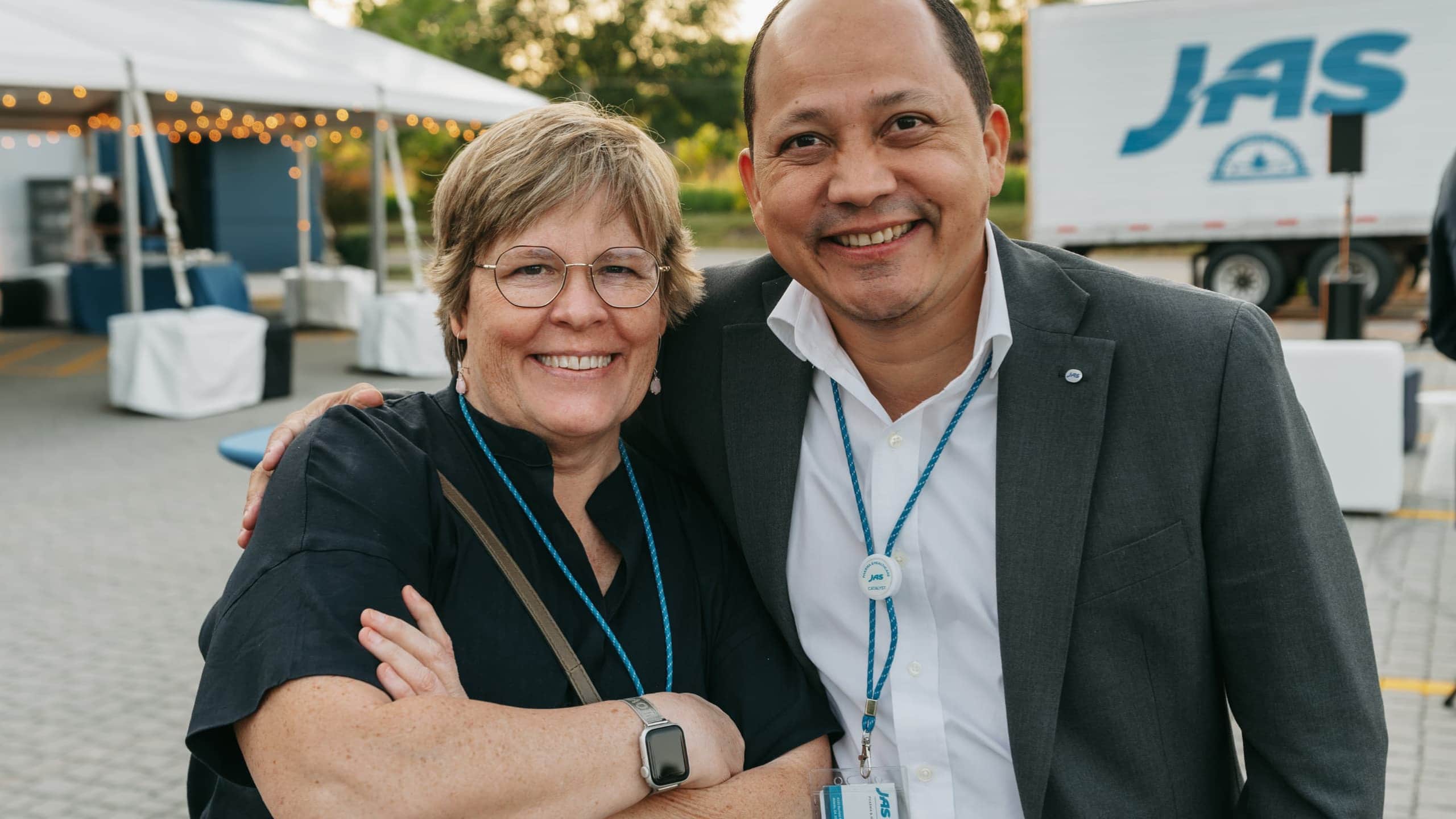 Two people smiling and standing together in an outdoor setting with a tent and truck in the background.