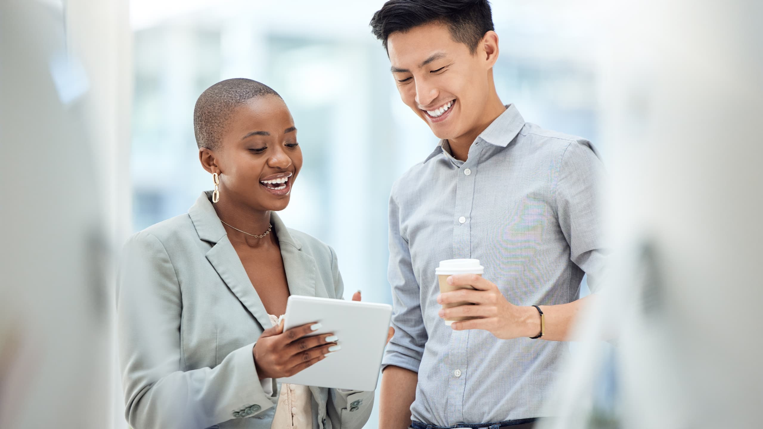 Two people smiling and looking at a tablet, one holding a coffee cup, in a bright indoor setting.