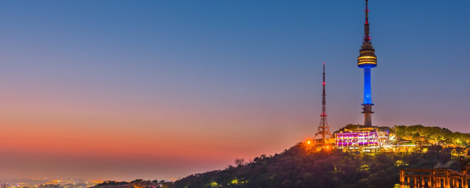 Cityscape with towers on a hill during sunset, featuring a colorful sky and illuminated buildings.