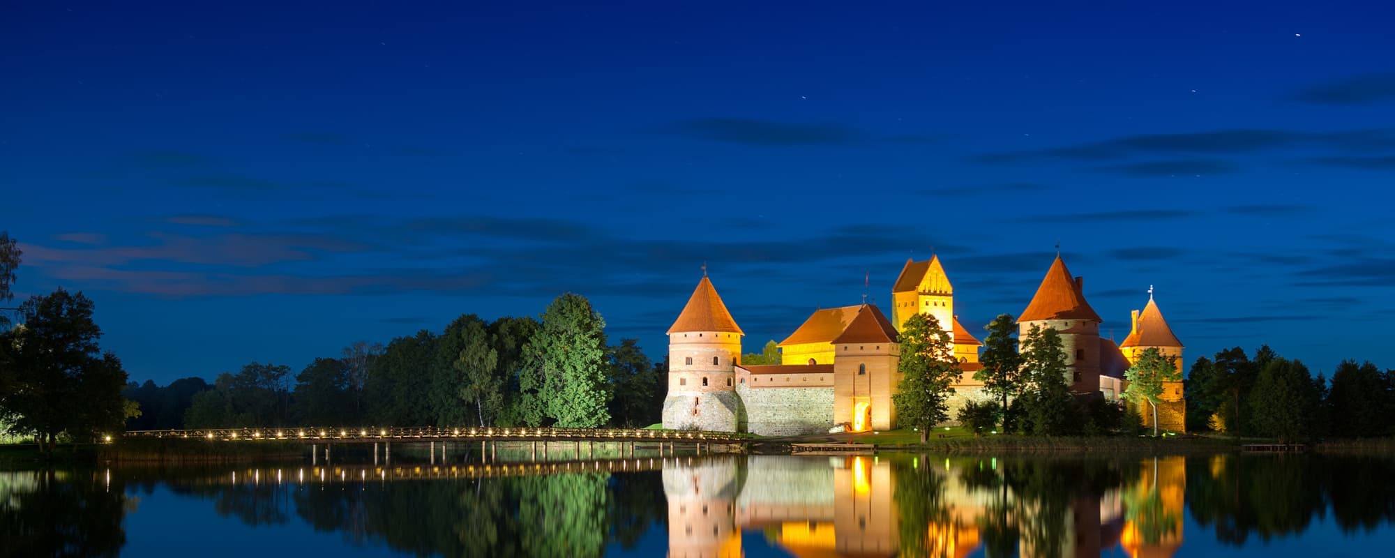 A brightly lit castle on an island with red-orange roofs reflects on a lake at dusk.