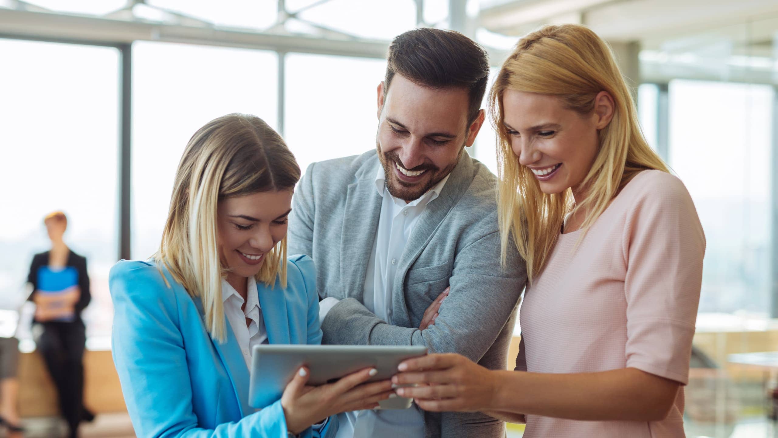 Three people smiling and looking at a tablet in a bright office setting.