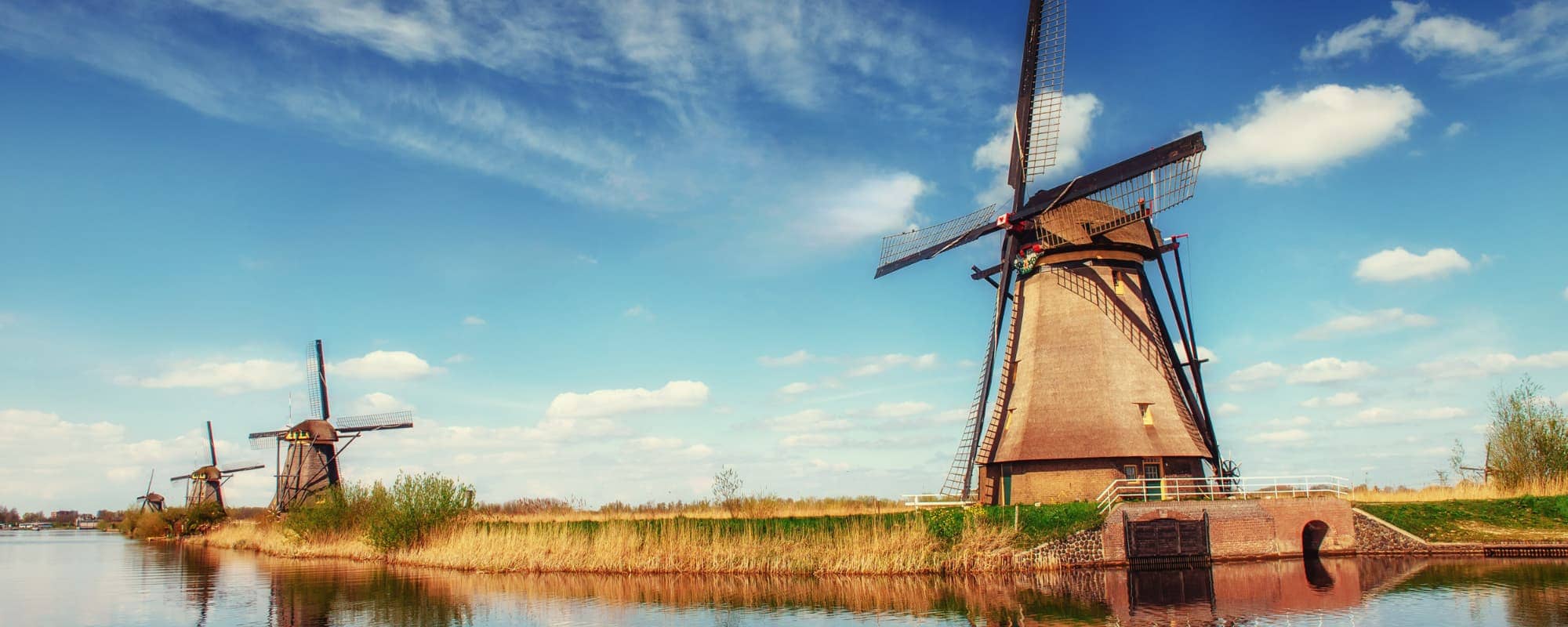 A rustic landscape with three traditional windmills beside a river under a bright blue sky with clouds.