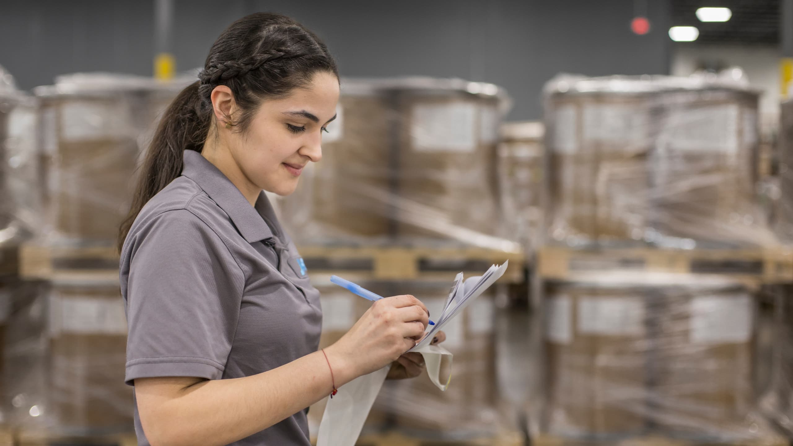 A woman writing on a clipboard in a warehouse, standing near stacked pallets.