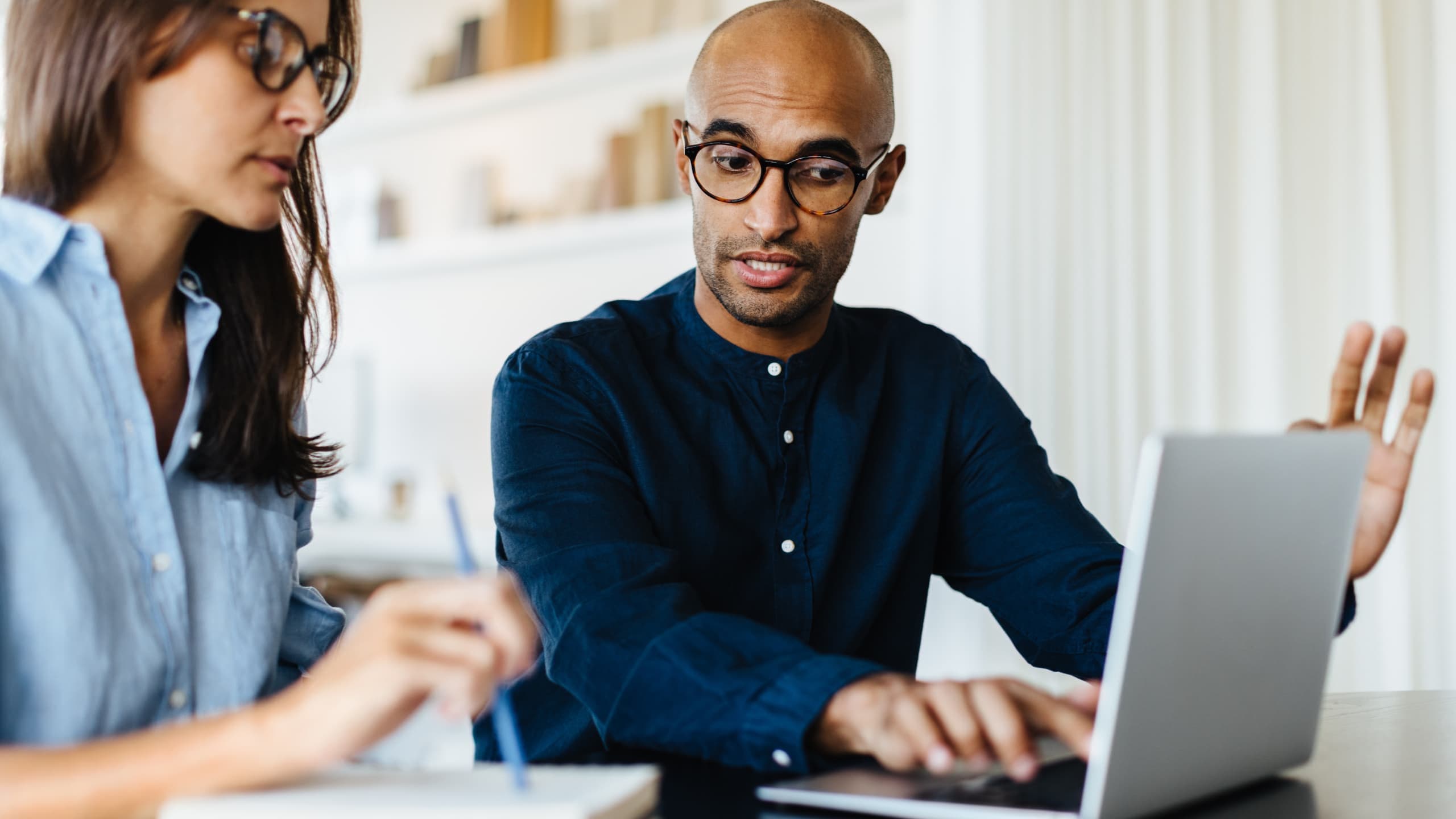 Two people in an office, one writing and the other talking while using a laptop.