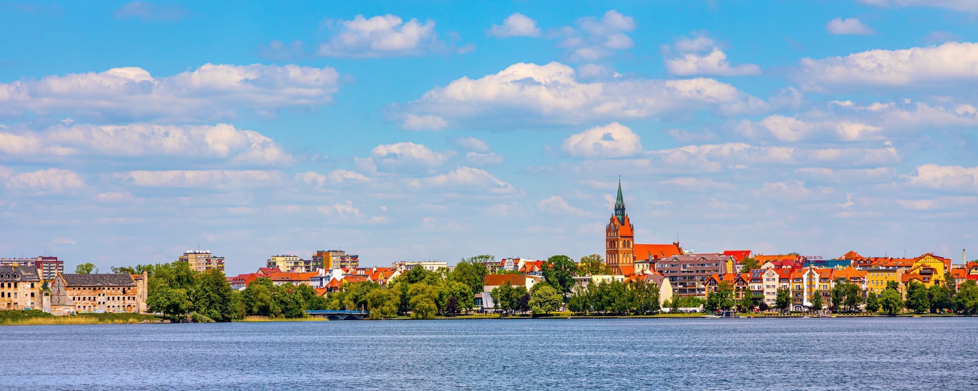 Panoramic view of a town with colorful buildings and a church spire by the water, under a blue sky with clouds.