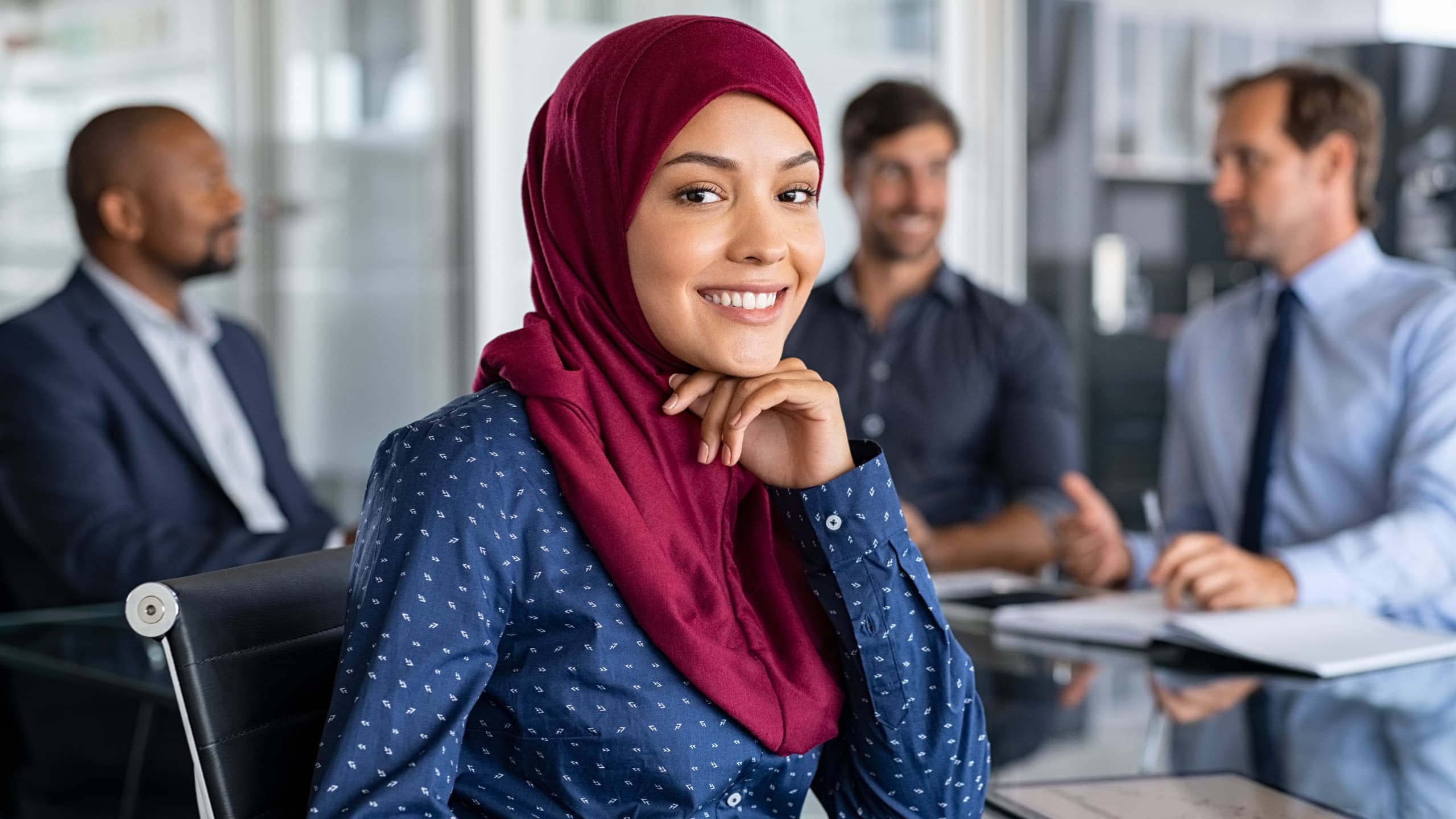 A woman in a maroon hijab and blue patterned shirt smiles at the camera in a conference room, with colleagues conversing in the background.