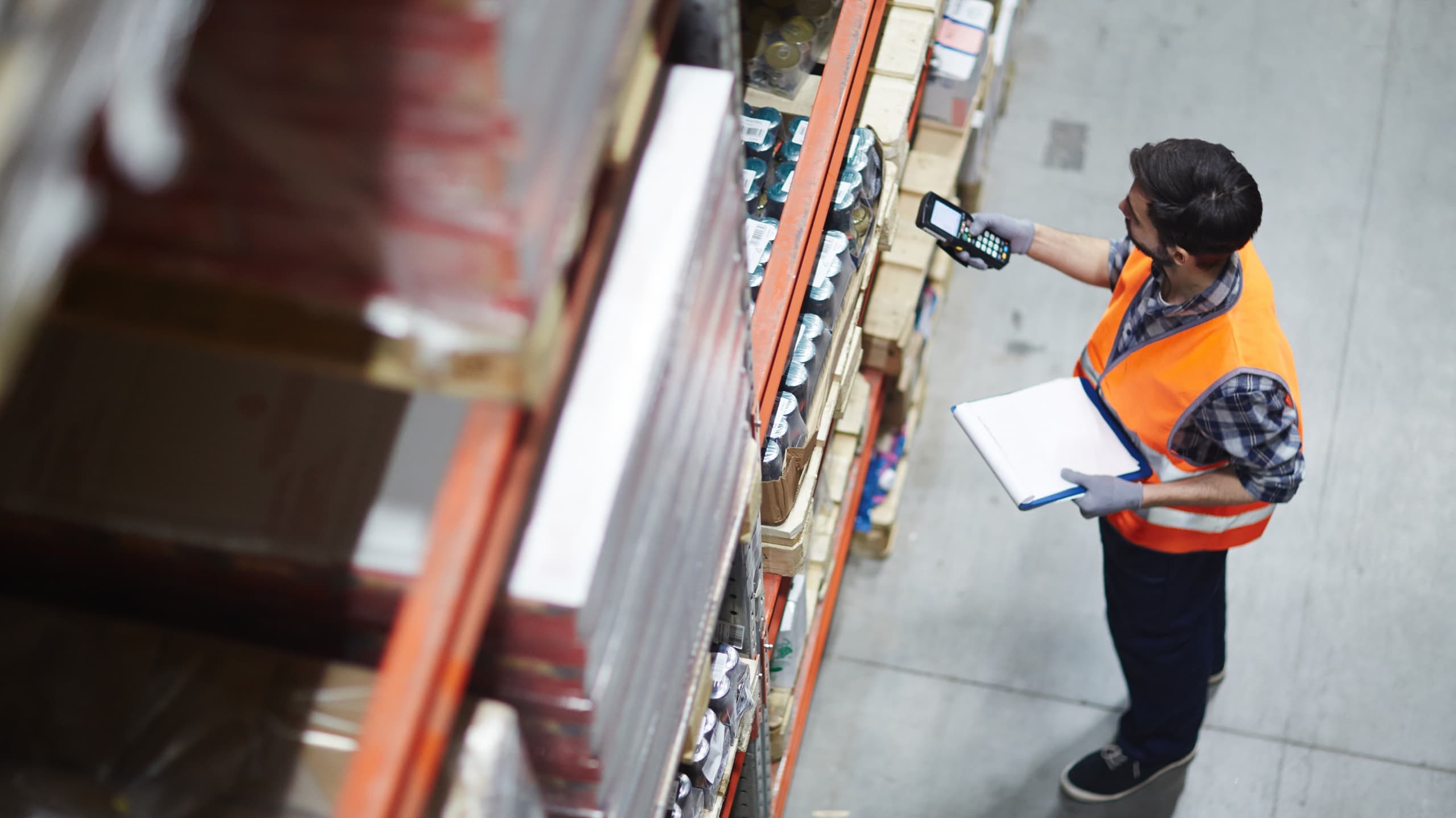 Warehouse worker in orange vest holding a device and clipboard in an aisle.
