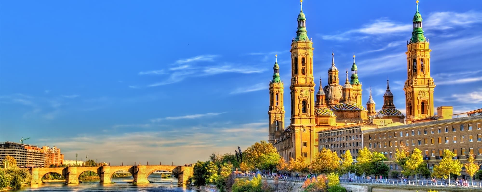 Panoramic view of a historic cathedral with tall spires, a stone bridge, and a river under a blue sky.