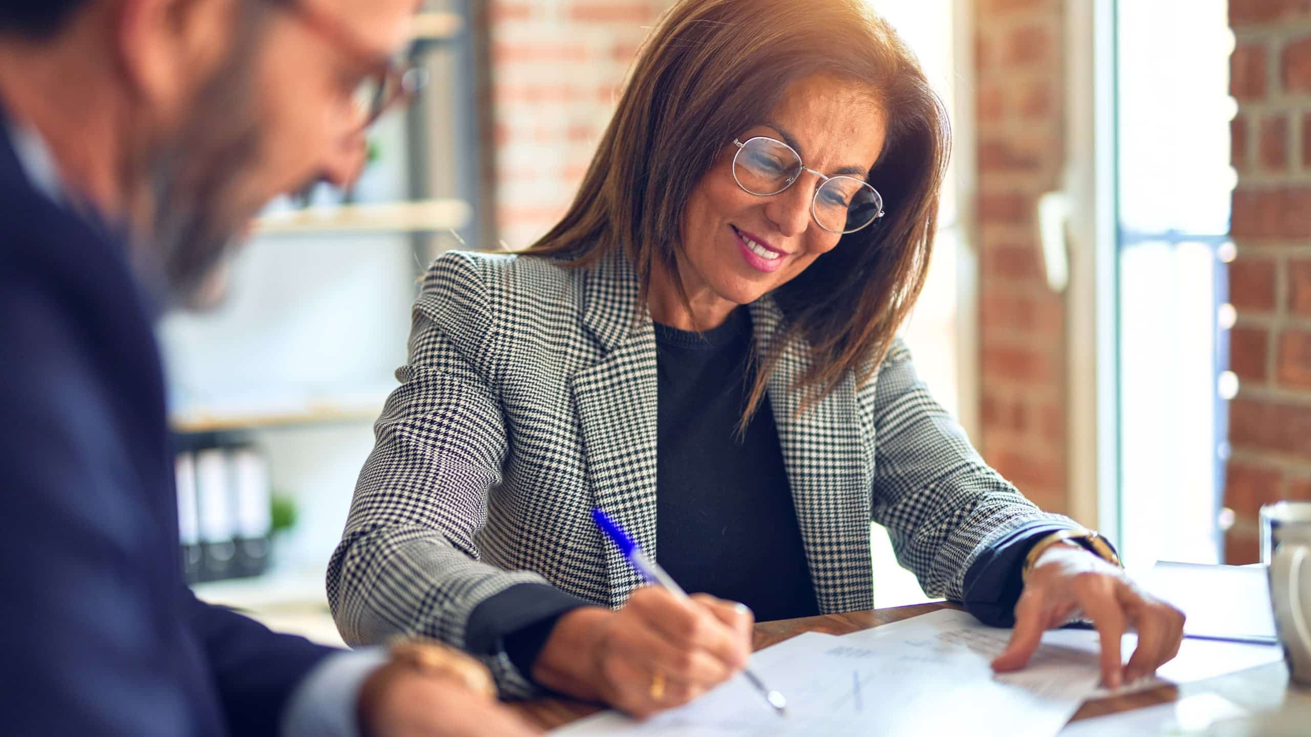 Woman in a checkered blazer writing with a pen at a table, next to a man in a suit.