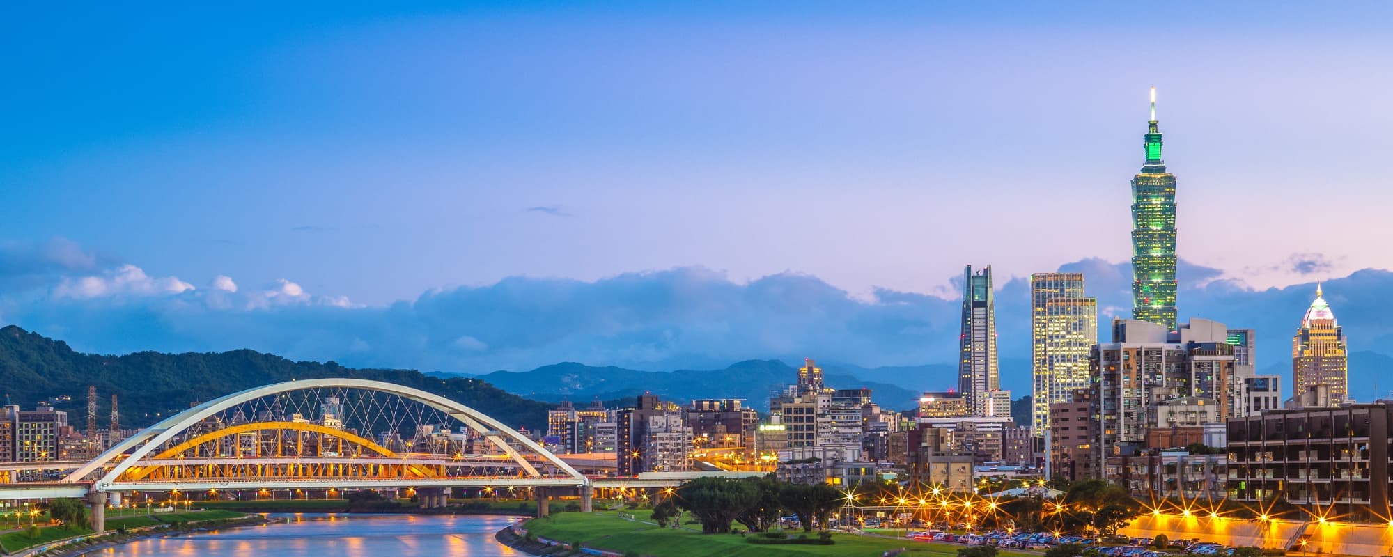 Cityscape at dusk with a skyscraper and a lit bridge over a river.