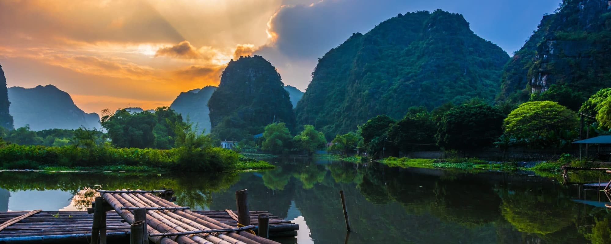 Bamboo dock over a calm lake with forested hills and sunset sky.