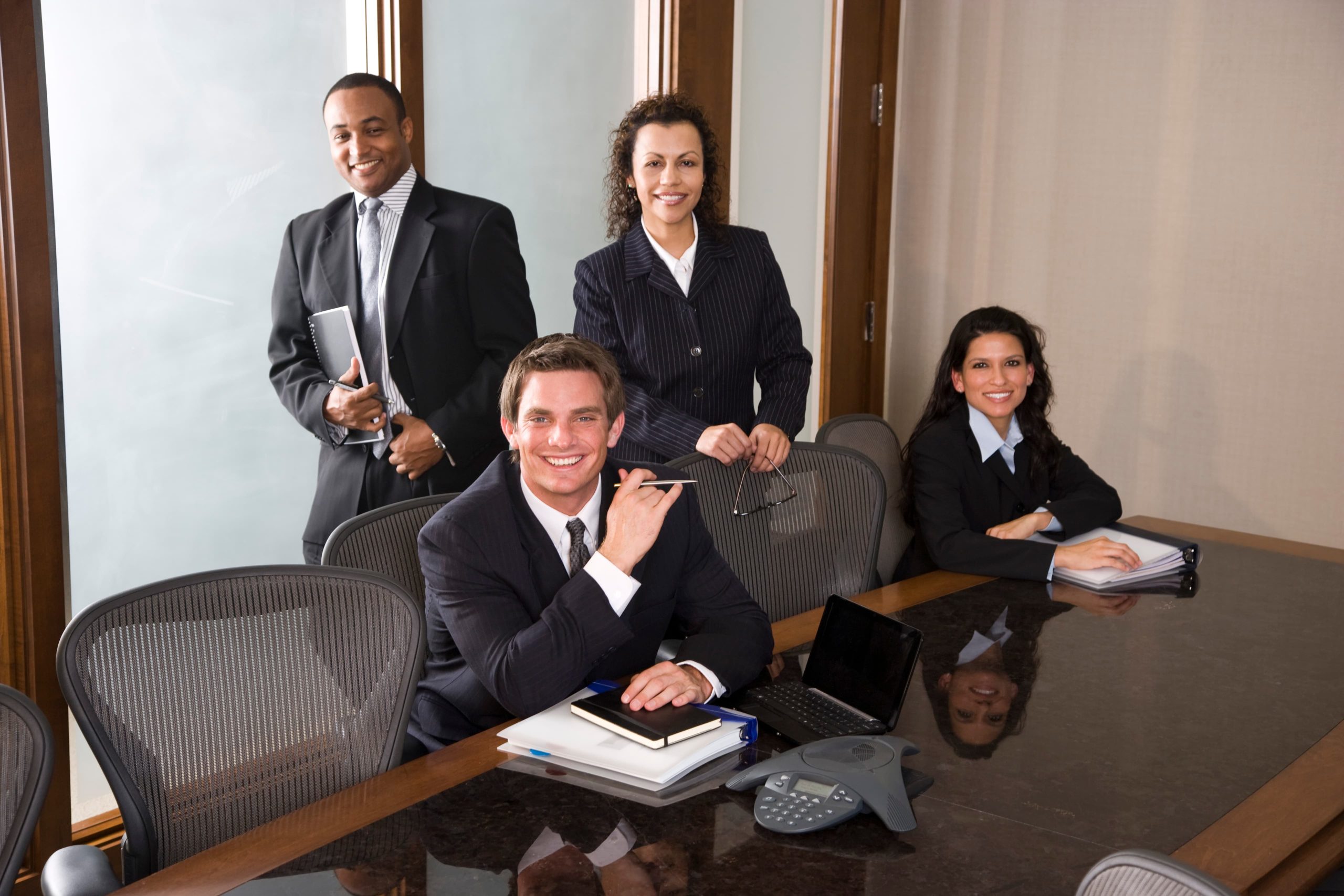Four professionals in business attire around a conference table with a laptop and conference phone.
