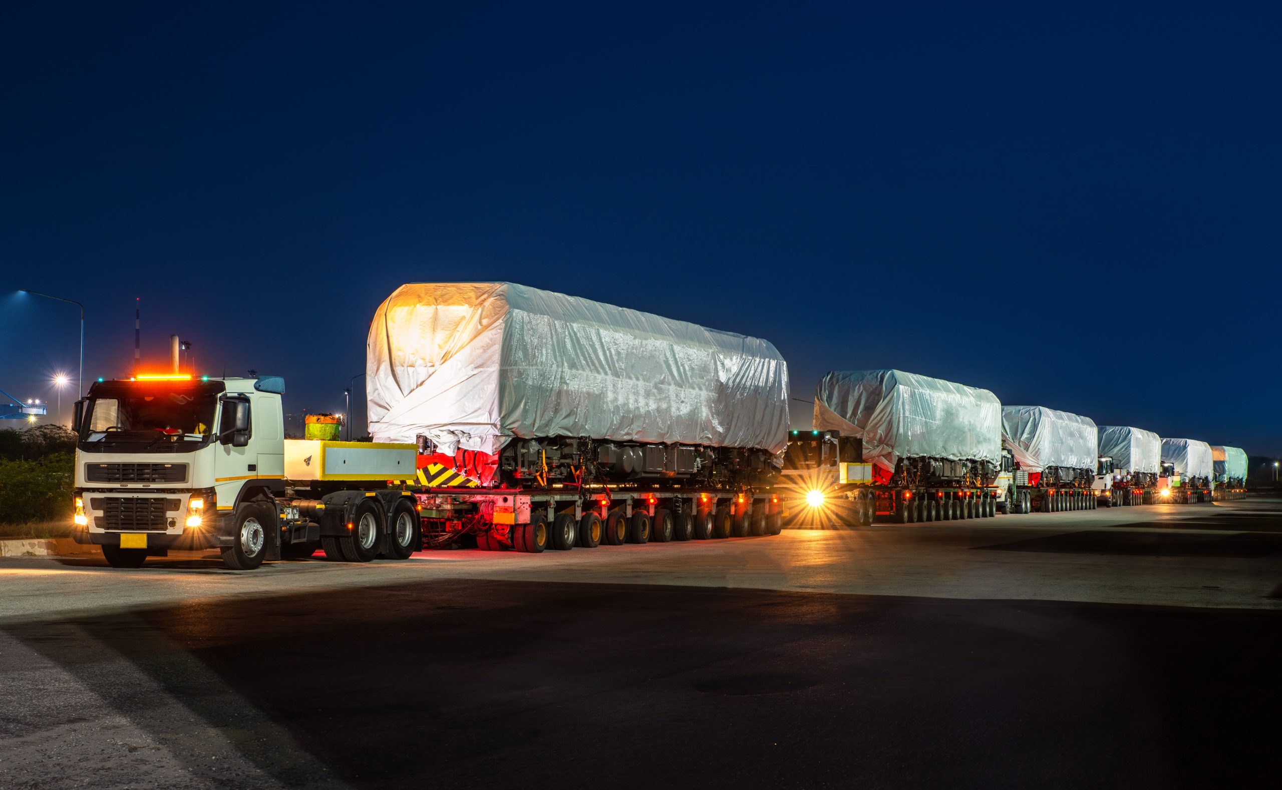 Convoy of large trucks carrying tarpaulin-covered loads at night, with headlights on.